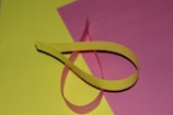 A yellow paper strip is twisted and placed on a flat surface with a pink background. The arrangement forms a loop, creating a simple, minimalistic view.