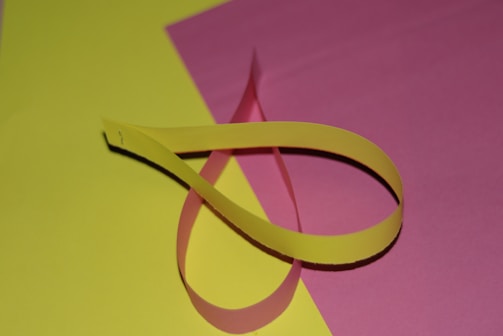 A yellow paper strip is twisted and placed on a flat surface with a pink background. The arrangement forms a loop, creating a simple, minimalistic view.