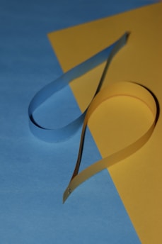 A paper craft composition featuring a strip of blue paper elegantly curled against a contrasting yellow background. The surfaces are joined at an oblique angle, creating a visually appealing intersection of shapes and shadows.