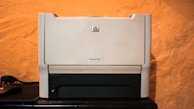 A beige laser printer sits on a dark surface against a textured orange wall. The printer has a compact design, featuring a small control panel and a paper tray visible at the bottom.