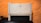 A beige laser printer sits on a dark surface against a textured orange wall. The printer has a compact design, featuring a small control panel and a paper tray visible at the bottom.