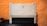 A beige laser printer sits on a dark surface against a textured orange wall. The printer has a compact design, featuring a small control panel and a paper tray visible at the bottom.