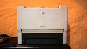 A beige laser printer sits on a dark surface against a textured orange wall. The printer has a compact design, featuring a small control panel and a paper tray visible at the bottom.