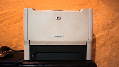 A beige laser printer sits on a dark surface against a textured orange wall. The printer has a compact design, featuring a small control panel and a paper tray visible at the bottom.