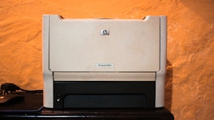 A beige laser printer sits on a dark surface against a textured orange wall. The printer has a compact design, featuring a small control panel and a paper tray visible at the bottom.