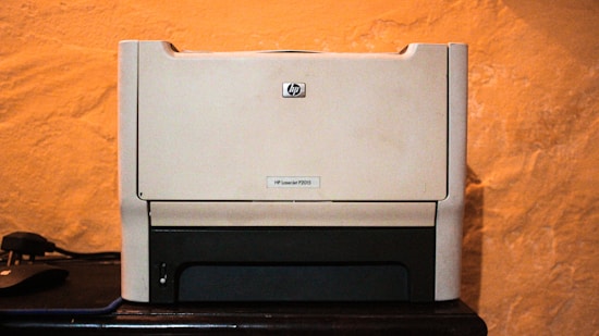 A beige laser printer sits on a dark surface against a textured orange wall. The printer has a compact design, featuring a small control panel and a paper tray visible at the bottom.