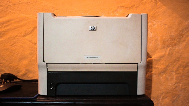 A beige laser printer sits on a dark surface against a textured orange wall. The printer has a compact design, featuring a small control panel and a paper tray visible at the bottom.