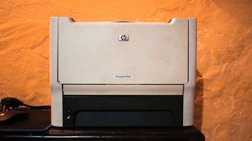 A beige laser printer sits on a dark surface against a textured orange wall. The printer has a compact design, featuring a small control panel and a paper tray visible at the bottom.