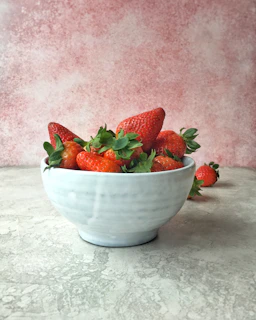 Strawberry powder in a small ceramic bowl next to fresh strawberries and leafy greens on a wooden surface.