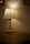 A table lamp with a pleated white shade stands on a wooden surface, casting a warm, diffused light against the beige wall. The lighting creates a cozy and intimate ambiance in the dimly lit space.