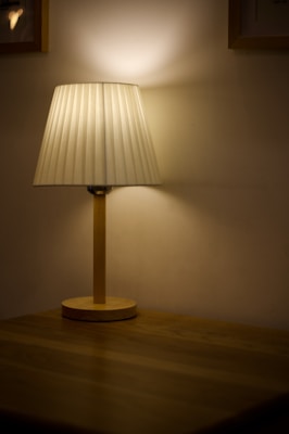 A table lamp with a pleated white shade stands on a wooden surface, casting a warm, diffused light against the beige wall. The lighting creates a cozy and intimate ambiance in the dimly lit space.