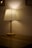 A table lamp with a pleated white shade stands on a wooden surface, casting a warm, diffused light against the beige wall. The lighting creates a cozy and intimate ambiance in the dimly lit space.