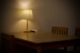 A wooden dining table is accompanied by wooden chairs in a dimly lit room. A lamp with a beige shade sits on the table, casting a soft, warm glow. There is a small white object on the table next to the lamp.