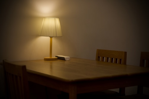 A wooden dining table is accompanied by wooden chairs in a dimly lit room. A lamp with a beige shade sits on the table, casting a soft, warm glow. There is a small white object on the table next to the lamp.