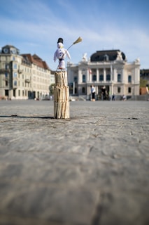 A small snowman figure stands atop a stack of wooden sticks in an open plaza. The background features historic buildings and an ornate theater, with a clear blue sky above.