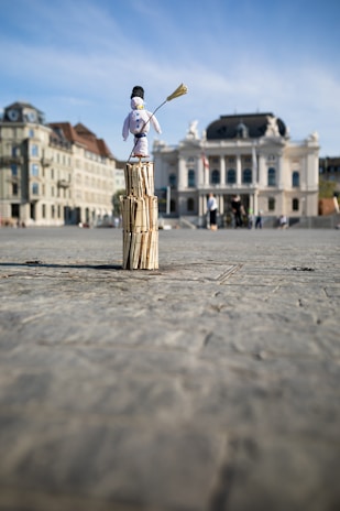 A small snowman figure stands atop a stack of wooden sticks in an open plaza. The background features historic buildings and an ornate theater, with a clear blue sky above.