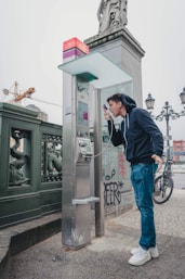 man in black jacket and blue denim jeans standing near black telephone booth during daytime