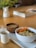A table setting featuring a white bowl filled with quinoa, topped with grated carrots, edamame, and seeds. Next to it is a wooden bowl of soup and a small white cup, all placed on a white tray. A glass of water and some reading material are also present. The background includes a blurred vase with greenery.