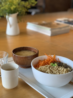 A table setting featuring a white bowl filled with quinoa, topped with grated carrots, edamame, and seeds. Next to it is a wooden bowl of soup and a small white cup, all placed on a white tray. A glass of water and some reading material are also present. The background includes a blurred vase with greenery.