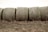 A row of large cylindrical hay bales lies on a dry grassy field. Each bale is tightly wrapped with netting and shows a mix of earthy tones. The ground beneath has sparse, dry grass, and the background is a light, overcast sky, giving the scene a rural and rustic feel.