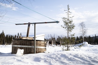 A snowy landscape featuring a wooden hot tub with a lid, surrounded by a partially snow-covered ground. In the background, two dark-colored horses stand near a fence, and evergreen trees are scattered throughout the scene. A tall, slender conifer tree and a simple clothesline structure appear in the foreground.