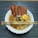 A bowl of instant noodles topped with slices of luncheon meat and a raw egg. The dish is garnished with greens and seasoning and is accompanied by a pair of decorative chopsticks resting on the bowl. The noodles are in a light broth, and the background features a striped placemat.