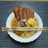 A bowl of instant noodles topped with slices of luncheon meat and a raw egg. The dish is garnished with greens and seasoning and is accompanied by a pair of decorative chopsticks resting on the bowl. The noodles are in a light broth, and the background features a striped placemat.