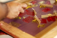 A hand is manipulating pieces of tape on a surface covered with red and yellow paint. The surface appears to be a glass or plastic sheet held in a wooden frame.