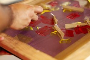 A hand is manipulating pieces of tape on a surface covered with red and yellow paint. The surface appears to be a glass or plastic sheet held in a wooden frame.