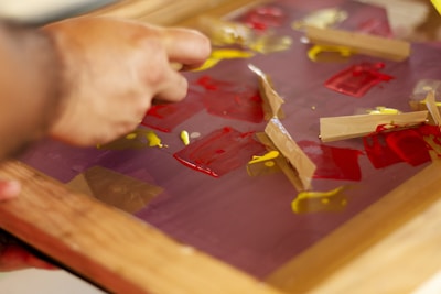 A hand is manipulating pieces of tape on a surface covered with red and yellow paint. The surface appears to be a glass or plastic sheet held in a wooden frame.