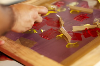 A hand is manipulating pieces of tape on a surface covered with red and yellow paint. The surface appears to be a glass or plastic sheet held in a wooden frame.