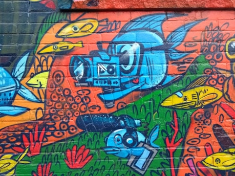 A colorful graffiti mural featuring a variety of stylized fish swimming through vibrant abstract patterns and backgrounds. The central focus is a large blue fish holding a camera, surrounded by smaller yellow fish and red coral-like designs.