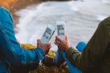 Two people sitting outdoors near the ocean, each holding a boxed water container with the text 'Boxed Water Is Better.' They are dressed in casual clothing, capturing a moment of relaxation and environmental consciousness.