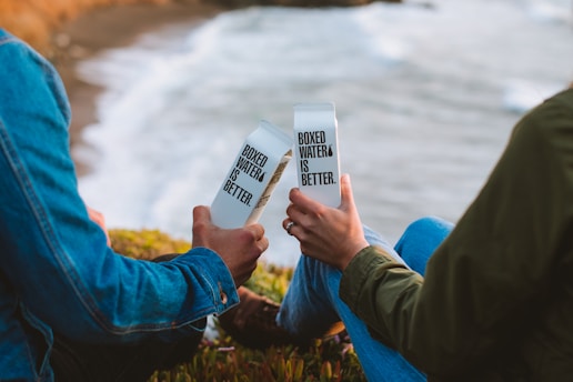 Two people sitting outdoors near the ocean, each holding a boxed water container with the text 'Boxed Water Is Better.' They are dressed in casual clothing, capturing a moment of relaxation and environmental consciousness.
