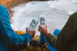 Two people sitting outdoors near the ocean, each holding a boxed water container with the text 'Boxed Water Is Better.' They are dressed in casual clothing, capturing a moment of relaxation and environmental consciousness.