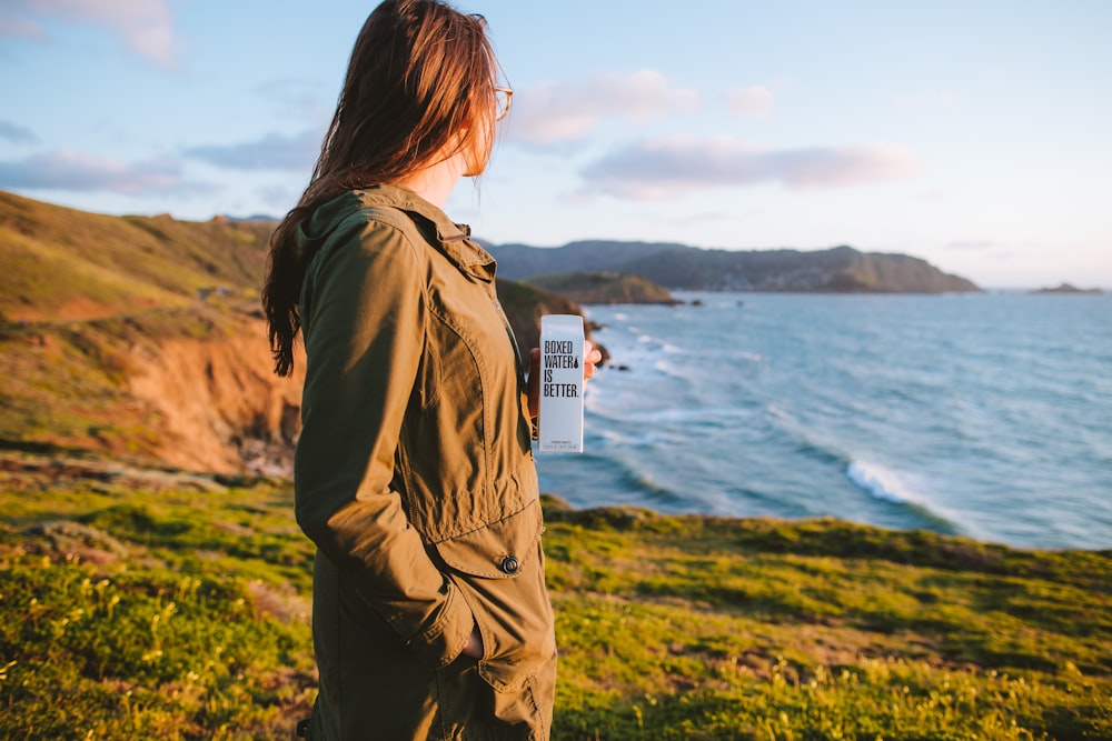 woman in brown jacket standing on green grass field near body of water during daytime