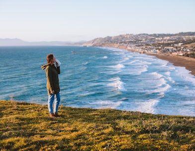 woman in black jacket and blue denim jeans standing on green grass field near body of on on on with