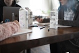 Three people are gathered around a table working, with notebooks and pens in front of them. Two cartons of boxed water are prominently displayed on the table. One carton has text saying 'Boxed Water Is Better' and is decorated with flowers and writing. A laptop, partially visible, is also on the table.