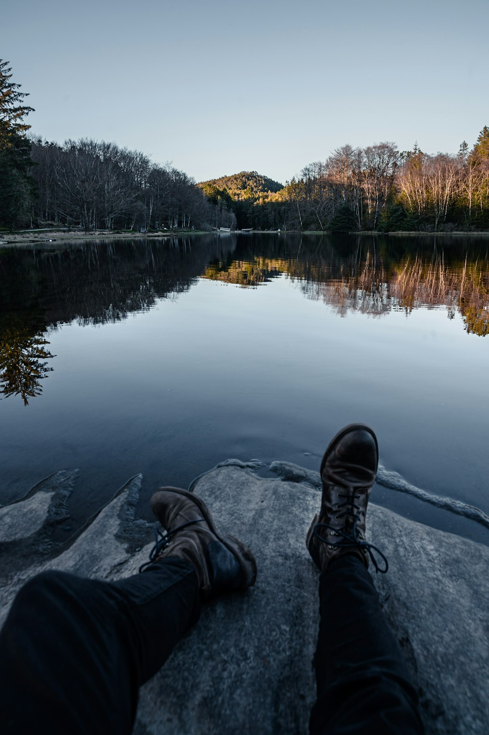 person in black pants and black shoes sitting on rock near lake during daytime