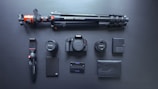 A neatly arranged set of photography equipment including a camera body, two camera lenses, a large tripod, a small handheld tripod, a camera battery, a battery charger, an external hard drive, a memory card holder, and a black leather wallet, all set against a dark surface.