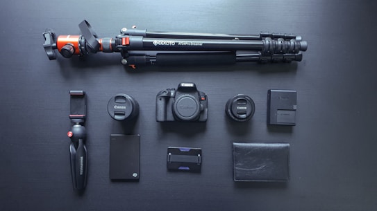 A neatly arranged set of photography equipment including a camera body, two camera lenses, a large tripod, a small handheld tripod, a camera battery, a battery charger, an external hard drive, a memory card holder, and a black leather wallet, all set against a dark surface.