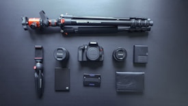 A neatly arranged set of photography equipment including a camera body, two camera lenses, a large tripod, a small handheld tripod, a camera battery, a battery charger, an external hard drive, a memory card holder, and a black leather wallet, all set against a dark surface.