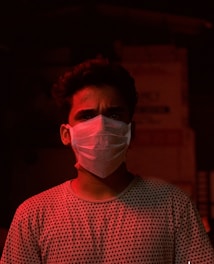 A person is wearing a white face mask and a polka dot shirt, illuminated by a red light in a dark environment. The background is indistinct and shadowy, enhancing the dramatic lighting effect on the person's face.