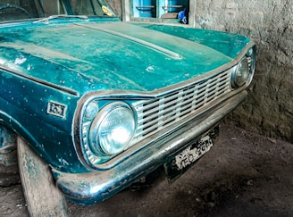 An old, vintage car with a teal blue paint job, showing signs of wear and dust accumulation. The car is parked in a dimly lit garage with rustic walls. The front bumper and headlights are prominently visible, and the license plate is weathered.