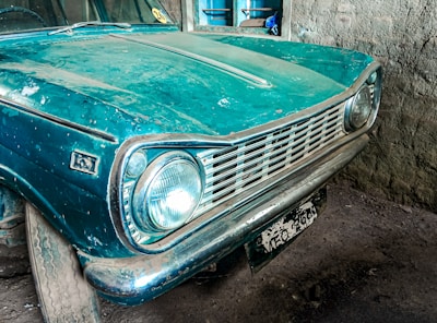 An old, vintage car with a teal blue paint job, showing signs of wear and dust accumulation. The car is parked in a dimly lit garage with rustic walls. The front bumper and headlights are prominently visible, and the license plate is weathered.