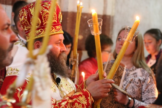A religious ceremony featuring a bearded man wearing a red and gold ornate robe and hat, holding large lit candles. Several people stand around him, including women, some holding candles as well. The scene is dimly lit, emphasizing the warm glow of the candlelight.
