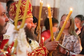 A religious ceremony featuring a bearded man wearing a red and gold ornate robe and hat, holding large lit candles. Several people stand around him, including women, some holding candles as well. The scene is dimly lit, emphasizing the warm glow of the candlelight.