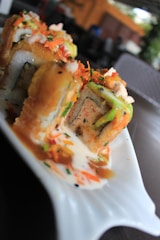 A close-up view of sushi rolls topped with colorful garnishes like shredded carrots and creamy sauce, presented on a unique wavy plate. The rolls are cut to reveal the intricate fillings inside, creating a vibrant and appetizing display.