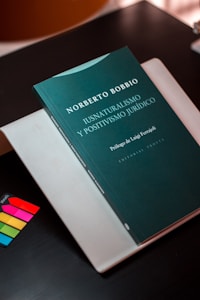 A book with a dark green cover is titled 'Iusnaturalismo y Positivismo Jurídico' by Norberto Bobbio is placed on top of a light-colored folder or book. There are colorful sticky note markers in pink, yellow, green, and blue on a dark surface next to the book.