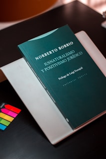 A book with a dark green cover is titled 'Iusnaturalismo y Positivismo Jur&iacute;dico' by Norberto Bobbio is placed on top of a light-colored folder or book. There are colorful sticky note markers in pink, yellow, green, and blue on a dark surface next to the book.